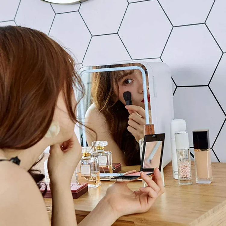 Woman using a white mini skincare fridge with mirror on makeup table.